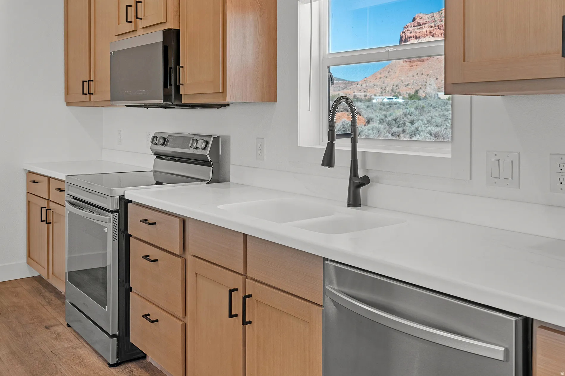 Modern kitchen featuring light wood cabinetry with dark hardware, white countertops, a double basin sink with a black gooseneck faucet, stainless steel appliances, and wood-finish flooring
