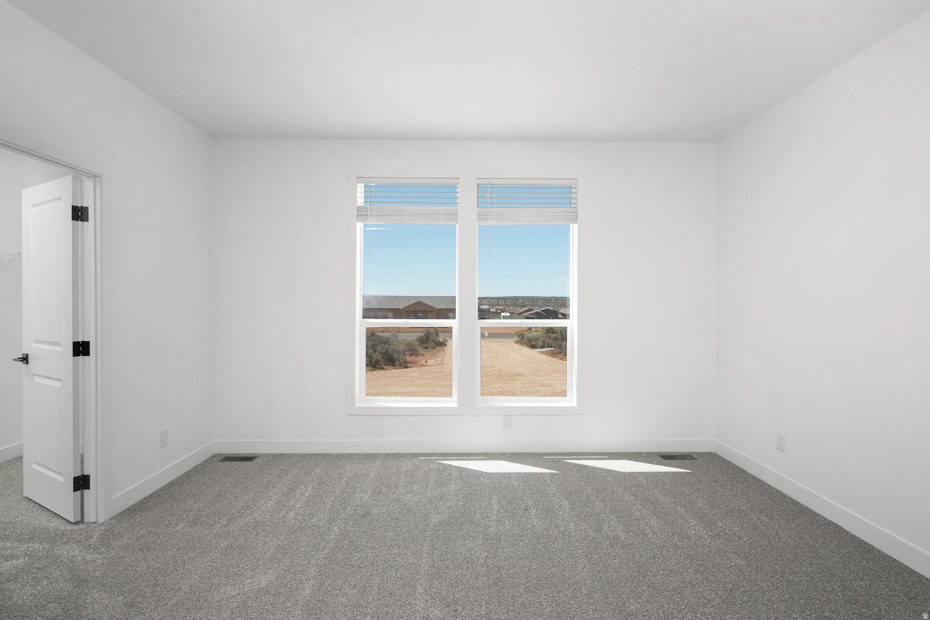 Bright room featuring a large window with horizontal blinds, gray carpet, white walls, and a white door with dark hardware