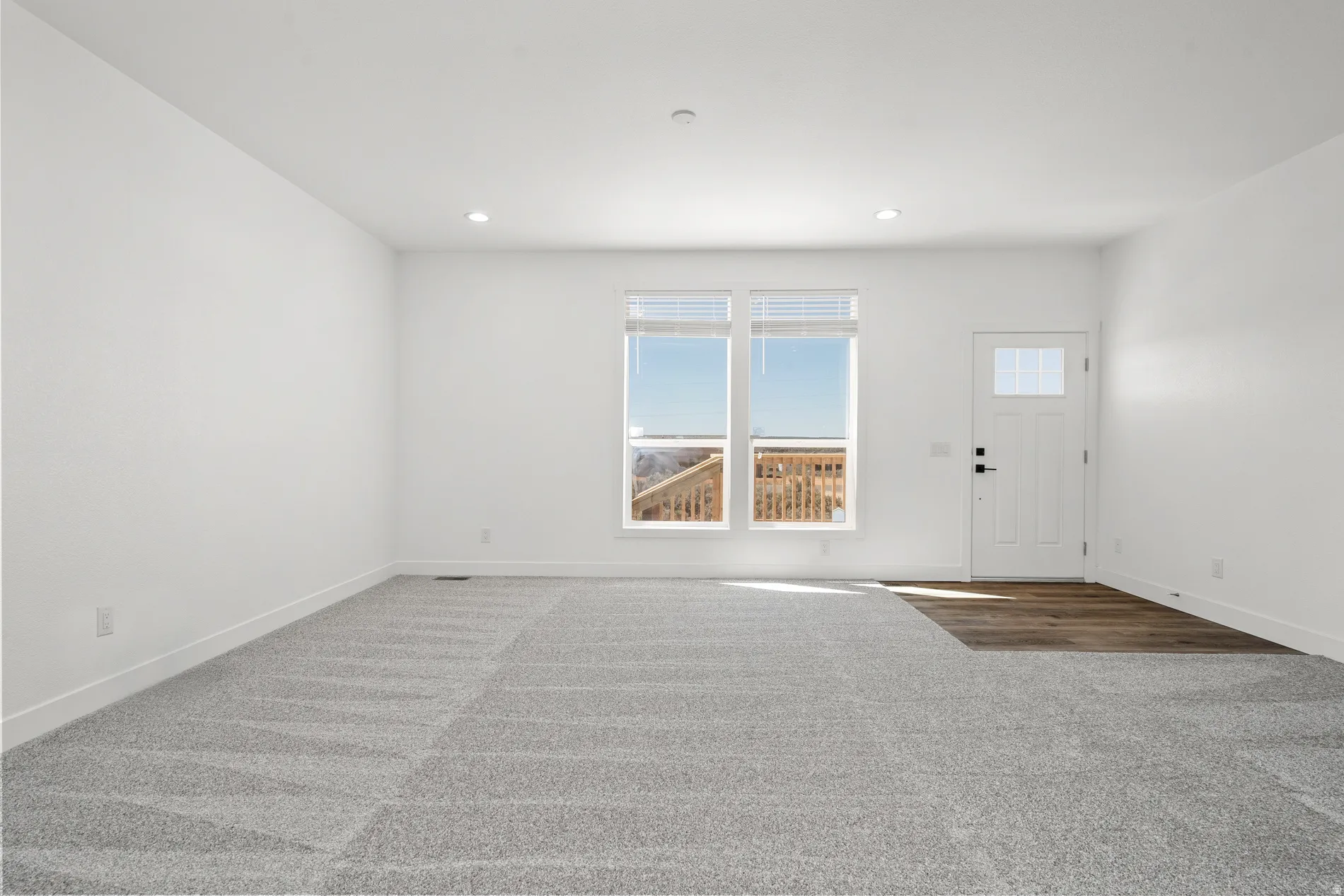 Spacious room featuring light gray carpet flooring transitioning to wood-finish flooring at the entryway