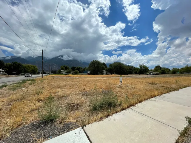 Expansive land parcel featuring a paved road, concrete sidewalk, distant mountainous scenery, and mature trees