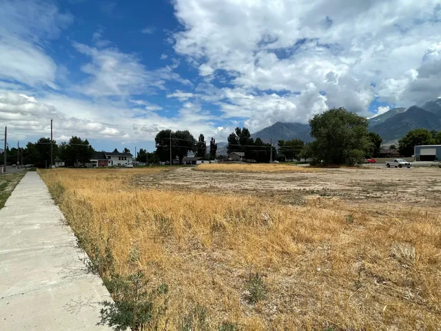 Expansive vacant lot featuring a paved sidewalk along one edge, mature trees in the background, and distant mountains