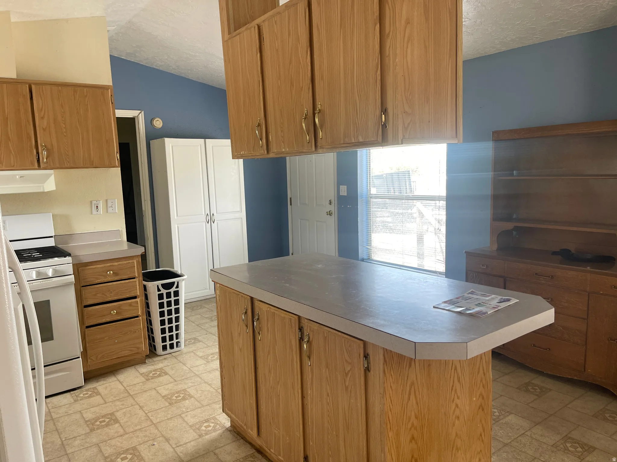 Kitchen featuring wood-finish cabinetry, a central island with a countertop, patterned vinyl flooring, a white range, and a built-in white pantry cabinet