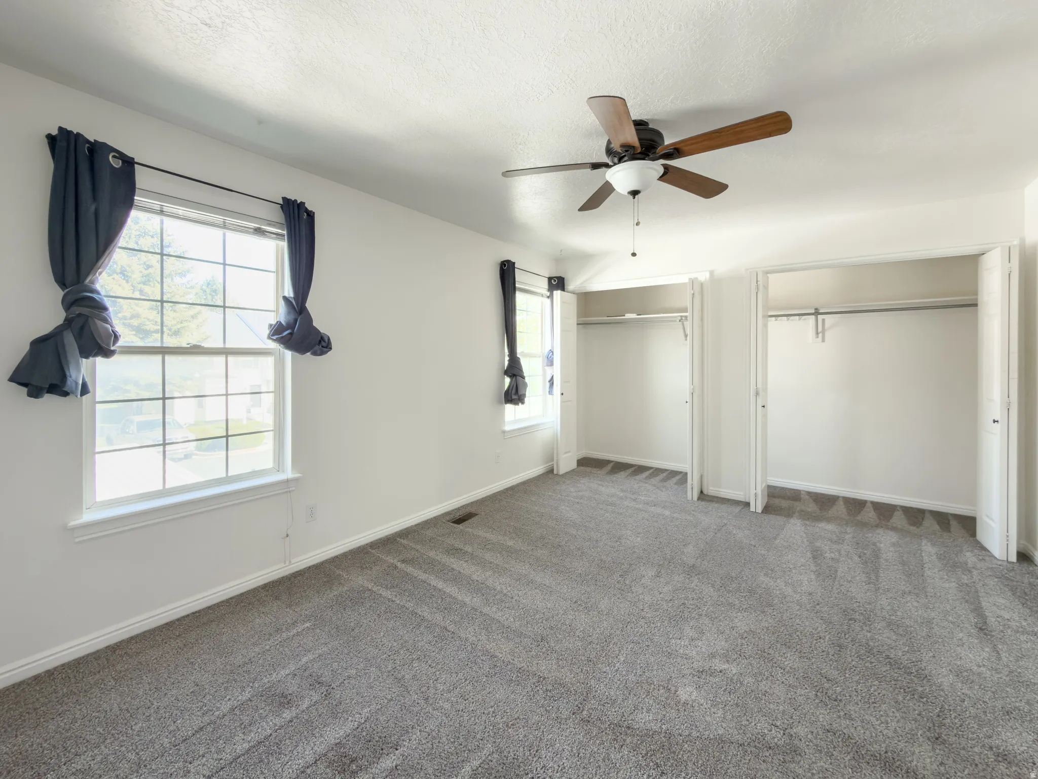 Bright interior space featuring a ceiling fan with wood-finish blades, two windows with white trim, and light gray carpeting