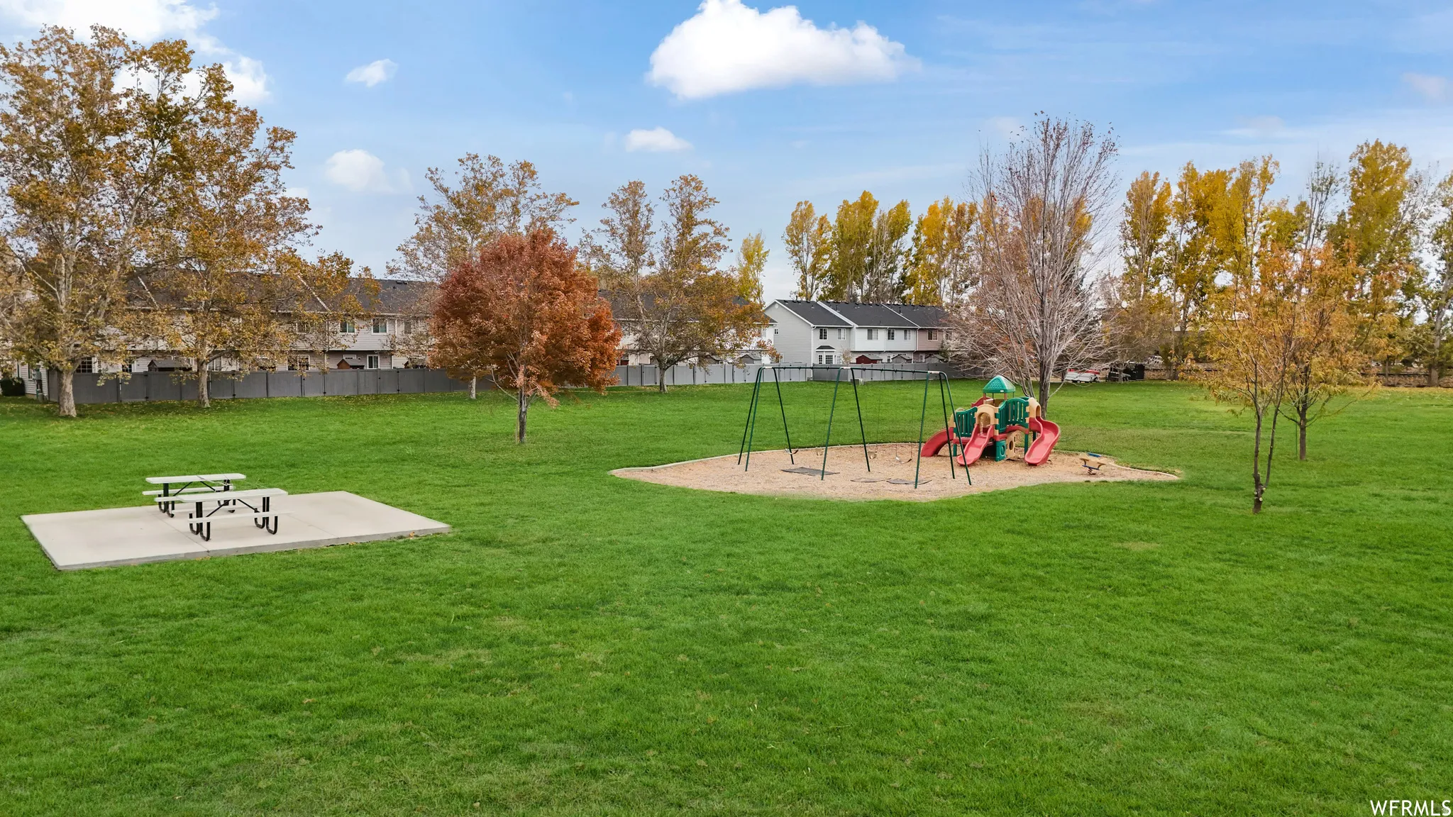 Patio area fence leads to this playground.Expansive green space featuring a playground with swings and slides, a picnic area with a concrete pad and picnic table, and mature trees with autumn foliage