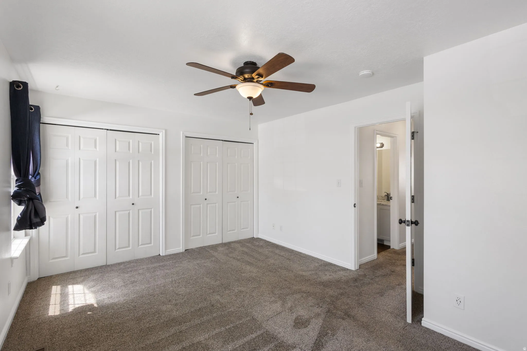 Carpeted interior space featuring a ceiling fan with integrated lighting, two sets of bi-fold closet doors, and a window with a deep sill