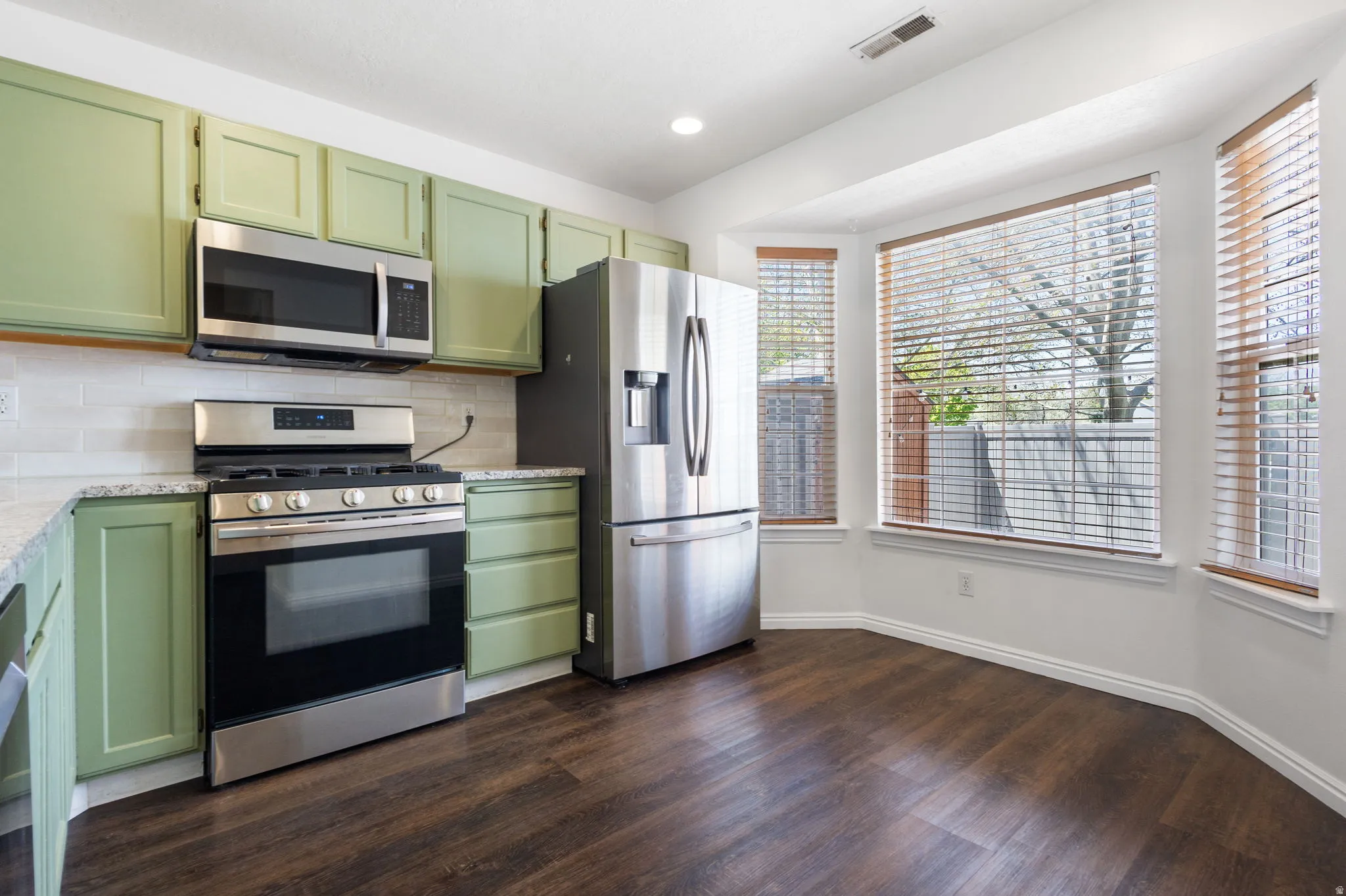 Kitchen featuring stainless steel appliances, light green cabinetry, light-toned backsplash, granite-style countertops, and wood-finish flooring