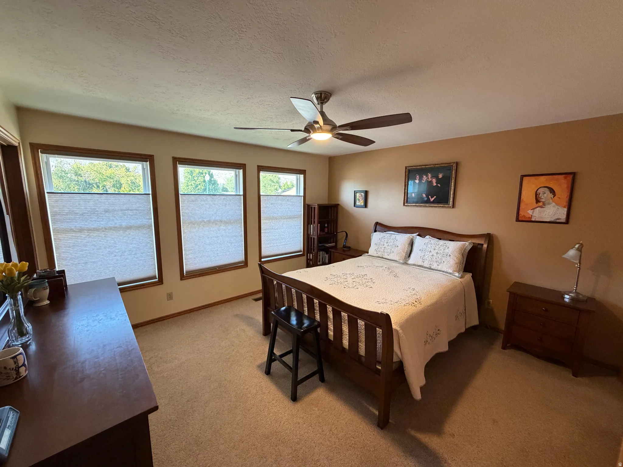 Carpeted bedroom featuring three expansive windows with privacy blinds, a ceiling fan with light fixture, and warm-toned wall paint