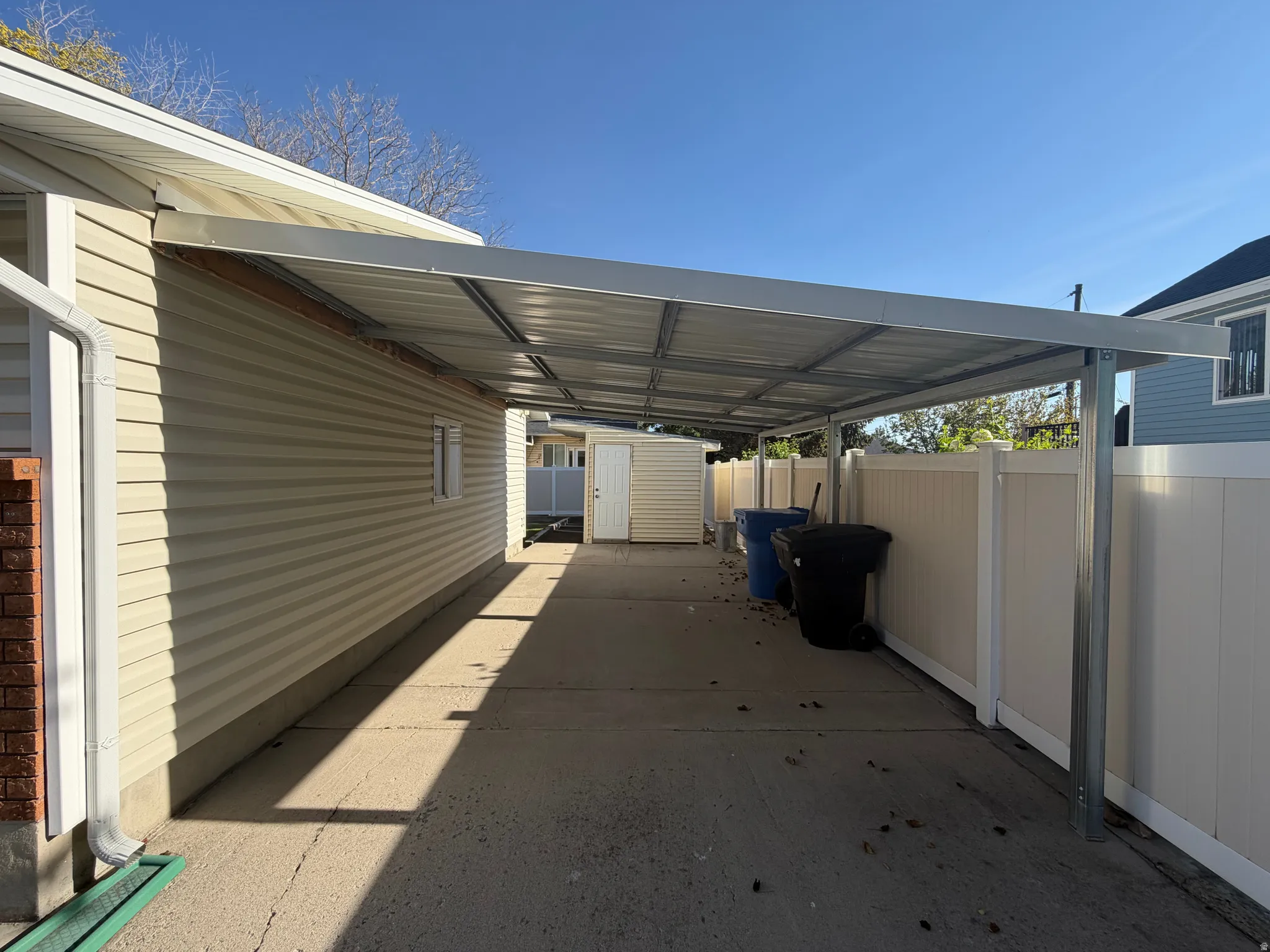 Covered concrete carport featuring a metal roof structure, tan vinyl siding, a white privacy fence, and an exterior storage shed
