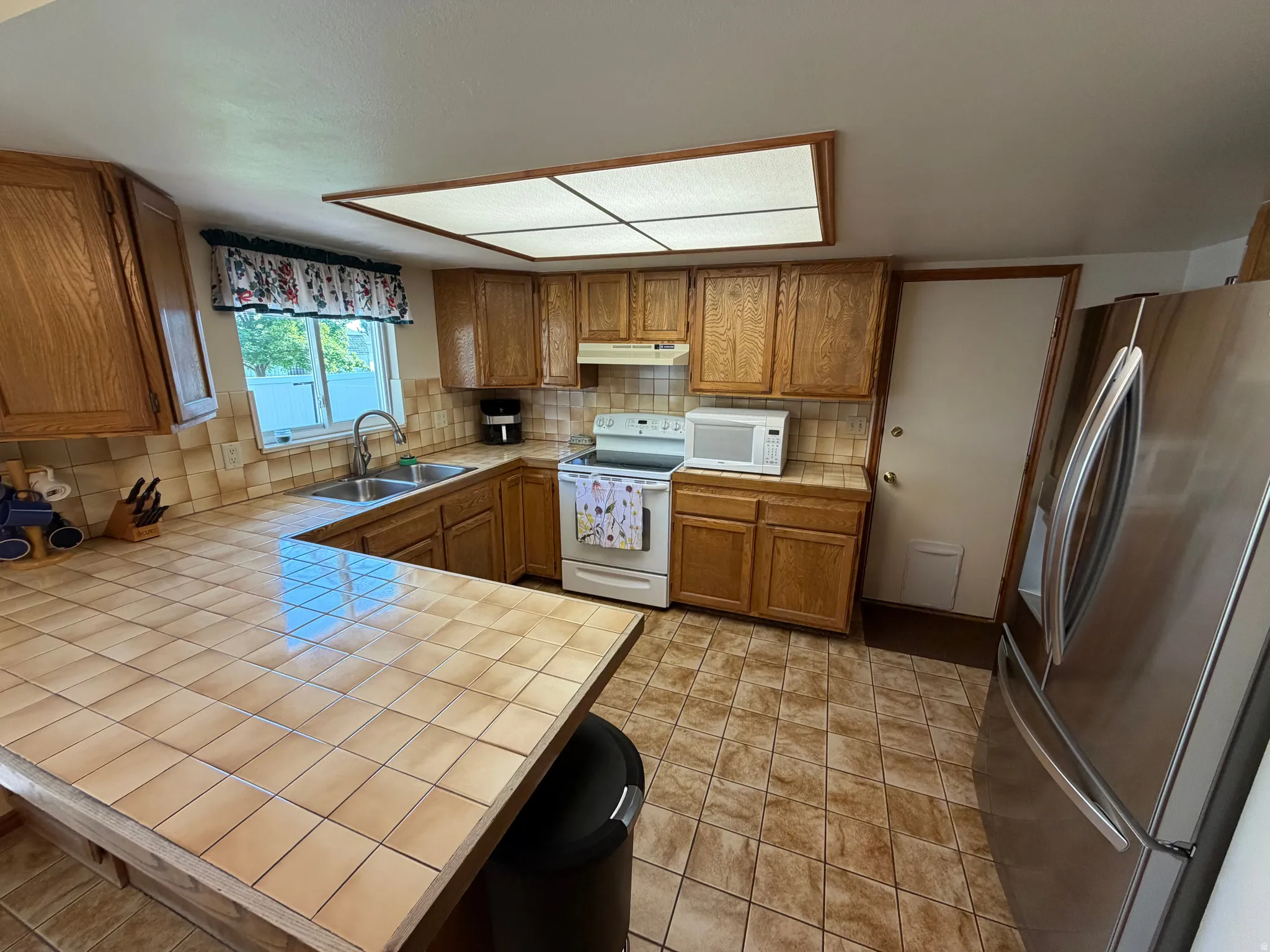 Kitchen featuring wood cabinetry, tile countertops, stainless steel double-door refrigerator, and a double basin sink