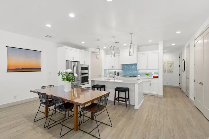 Open-concept kitchen and dining area featuring light wood-finish flooring, white cabinetry with contemporary hardware, stainless steel appliances, a blue subway tile backsplash, and recessed lighting