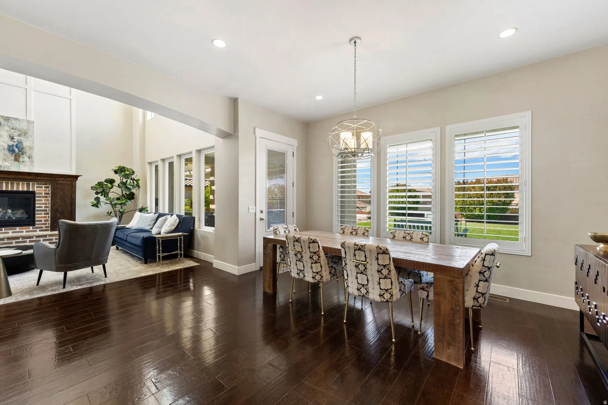 Open-concept dining area with dark wood-finish flooring, a contemporary chandelier, and three large windows featuring plantation shutters