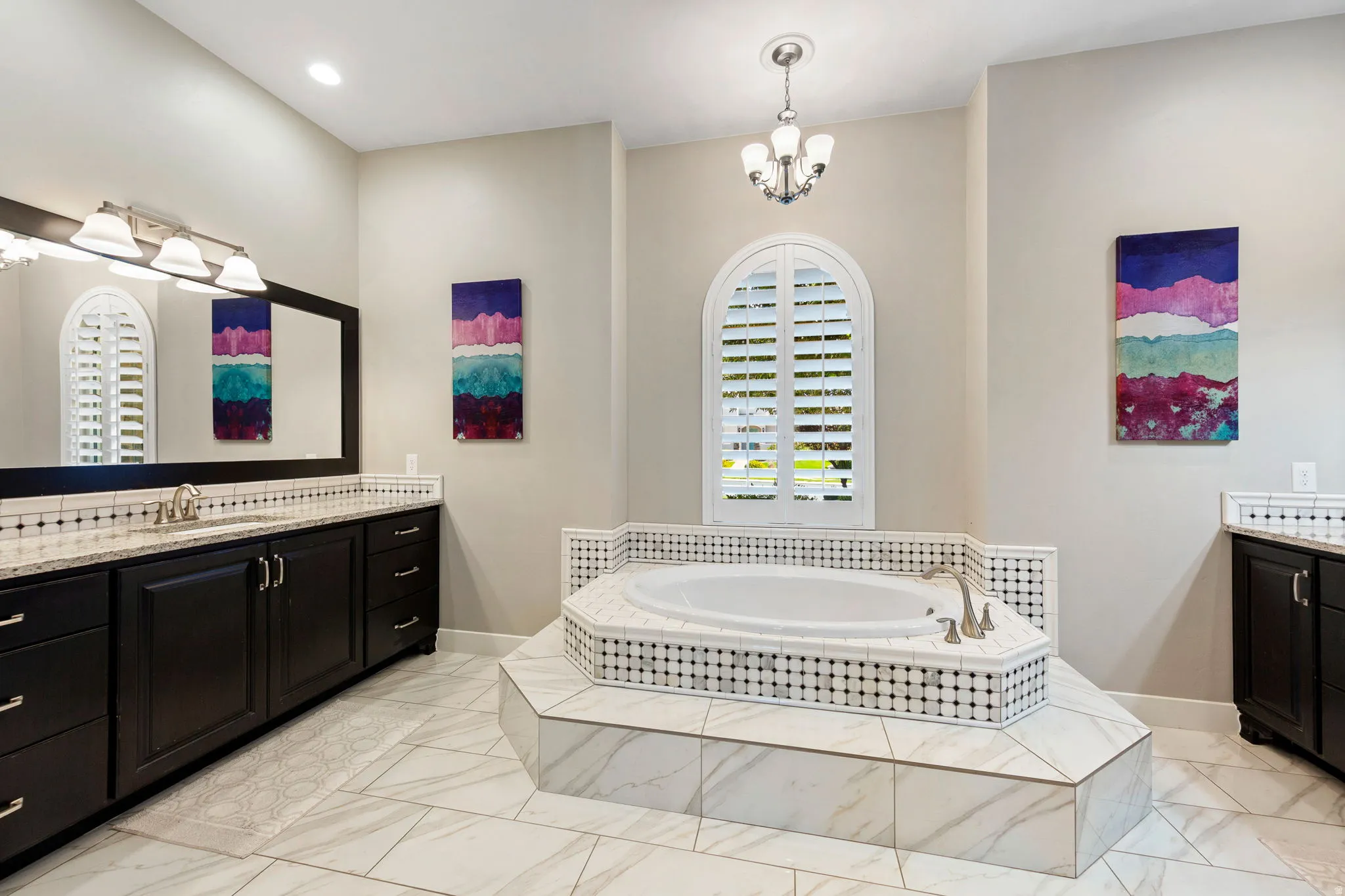 Spacious bathroom featuring a built-in corner soaking tub, arched window with plantation shutters, dual vanity with dark wood cabinetry, white countertops, and tile flooring