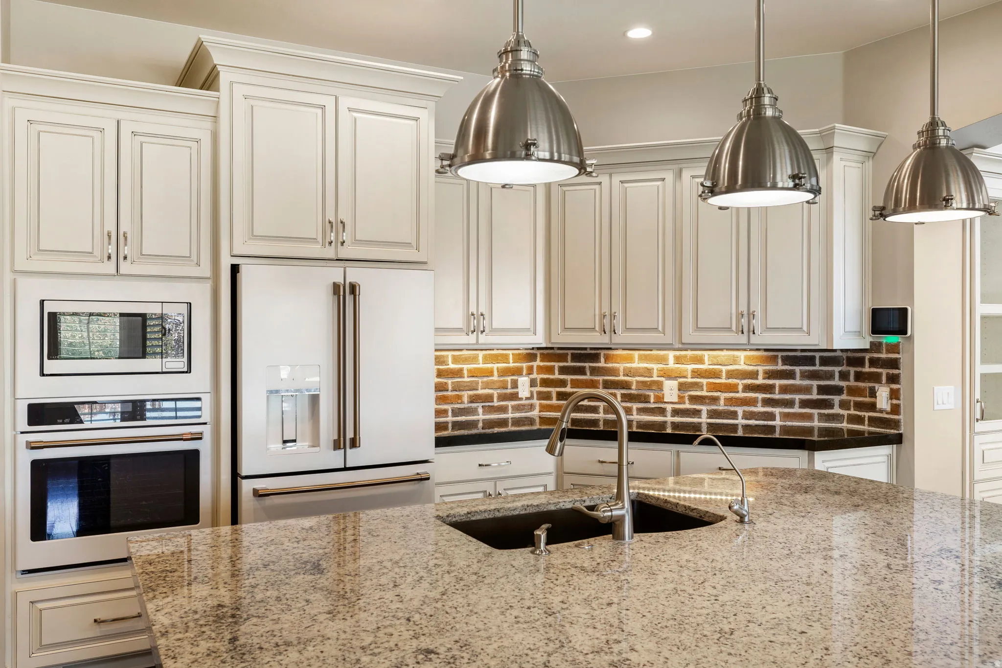Kitchen featuring a multi-tone granite island with an undermount sink, three industrial-style pendant lights, an integrated microwave and oven, a side-by-side refrigerator, and a brick-patterned backsplash