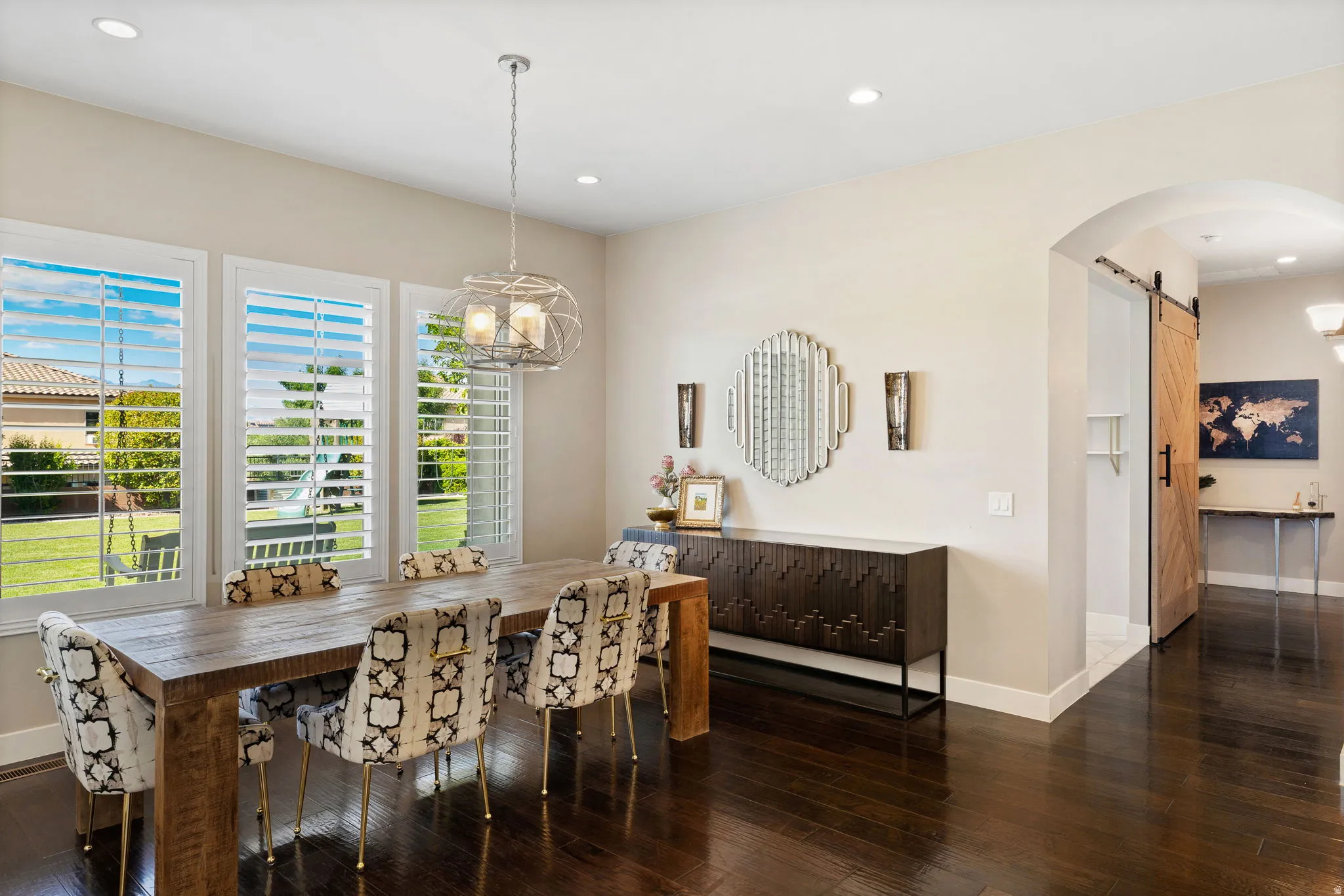 Dining area featuring dark wood-finish flooring, three large windows with white louvered shutters, and a modern geometric pendant light fixture