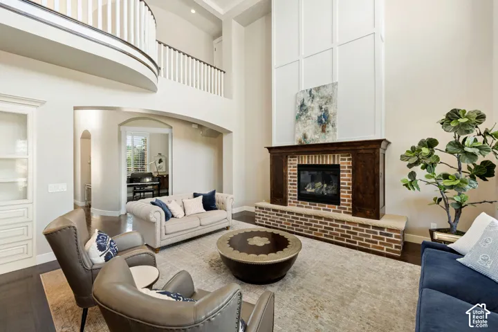 Two-story living area featuring a brick and wood-finish fireplace, rich wood-finish flooring, a curved balcony with white balusters, and an arched entryway