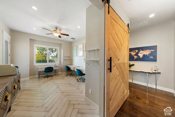 Natural wood barn door with black hardware opening to a room featuring light wood-finish flooring in a herringbone pattern, recessed lighting, a ceiling fan, and a window with plantation shutters