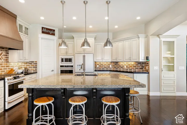 Spacious kitchen featuring a large island with a stone-finish countertop, white cabinetry, a brick-style backsplash, stainless steel appliances, and dark wood-finish flooring