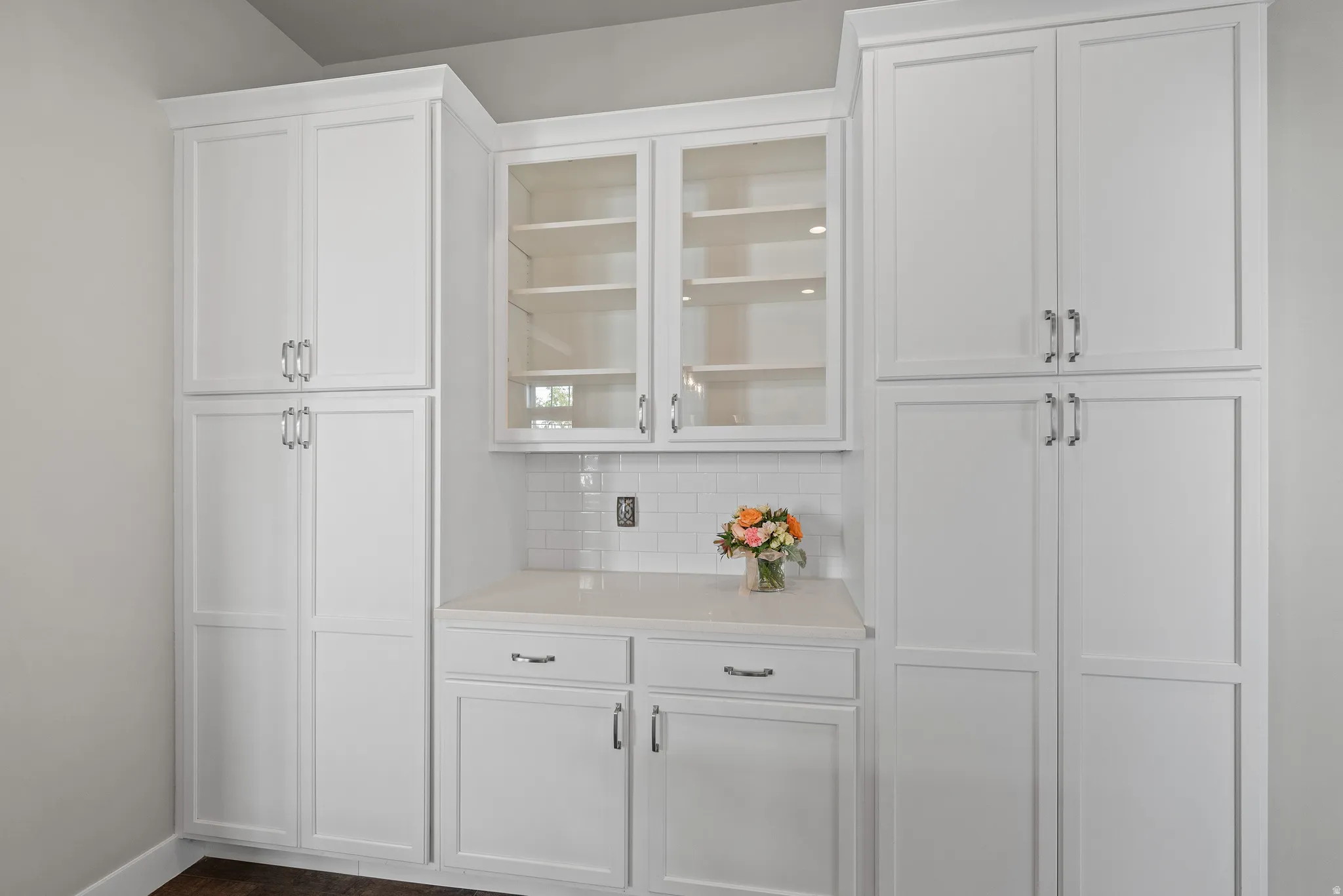White shaker cabinetry with crown molding, glass-front upper cabinets, light-toned countertop, and subway tile backsplash