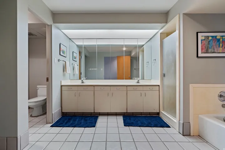 Spacious bathroom featuring a double vanity with ample counter space, extensive mirror cabinetry, a glass-enclosed shower, a separate commode area, and white tile flooring