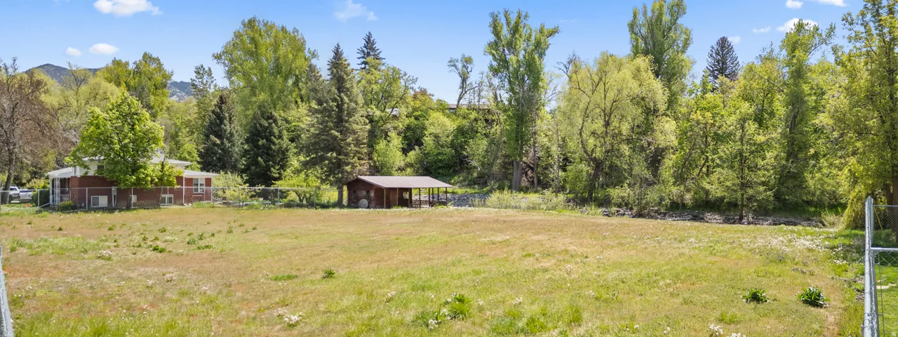 Expansive open lot featuring a detached outbuilding with covered space, surrounded by mature trees and natural landscaping