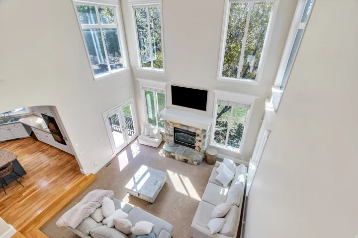 Two-story living area featuring a stone fireplace, large windows, French doors, carpet flooring, and an open concept to the kitchen with wood-finish flooring