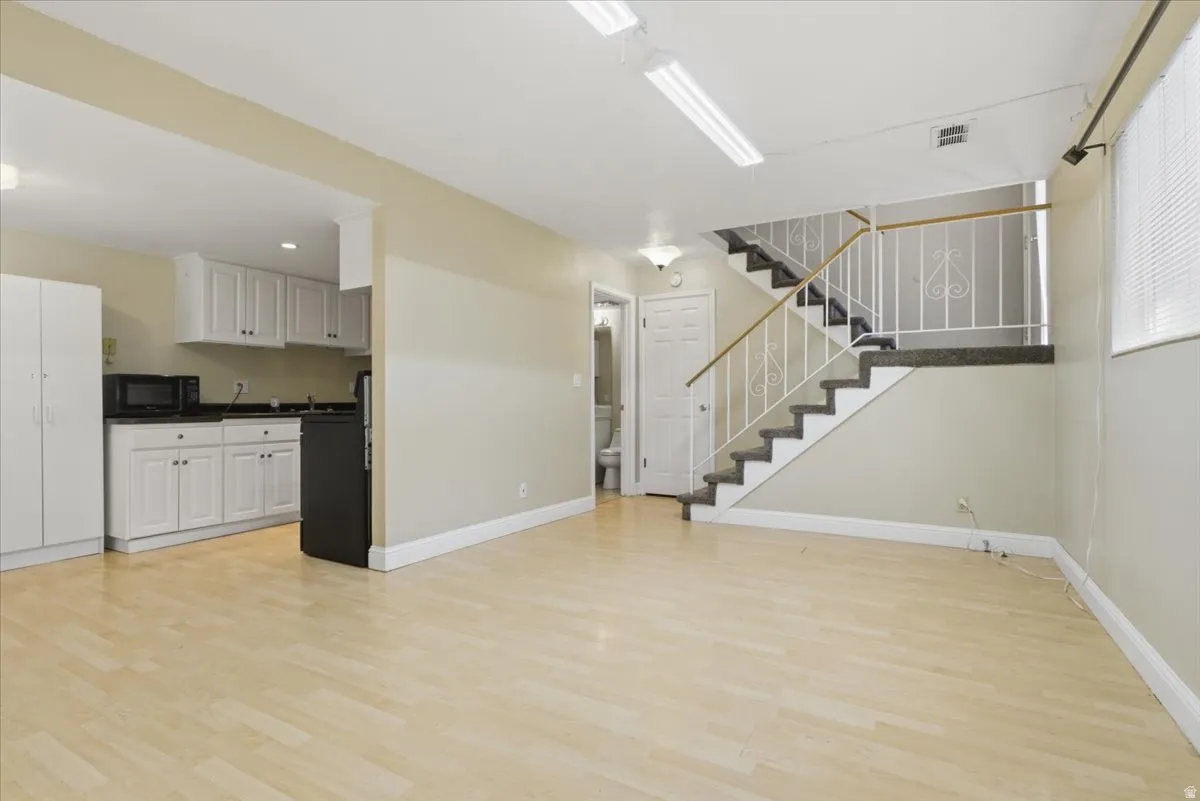 Open-concept living space featuring light wood-finish flooring, a contemporary staircase with ornamental railing, a compact kitchen with white cabinetry and dark countertops, recessed lighting, and a window with blinds