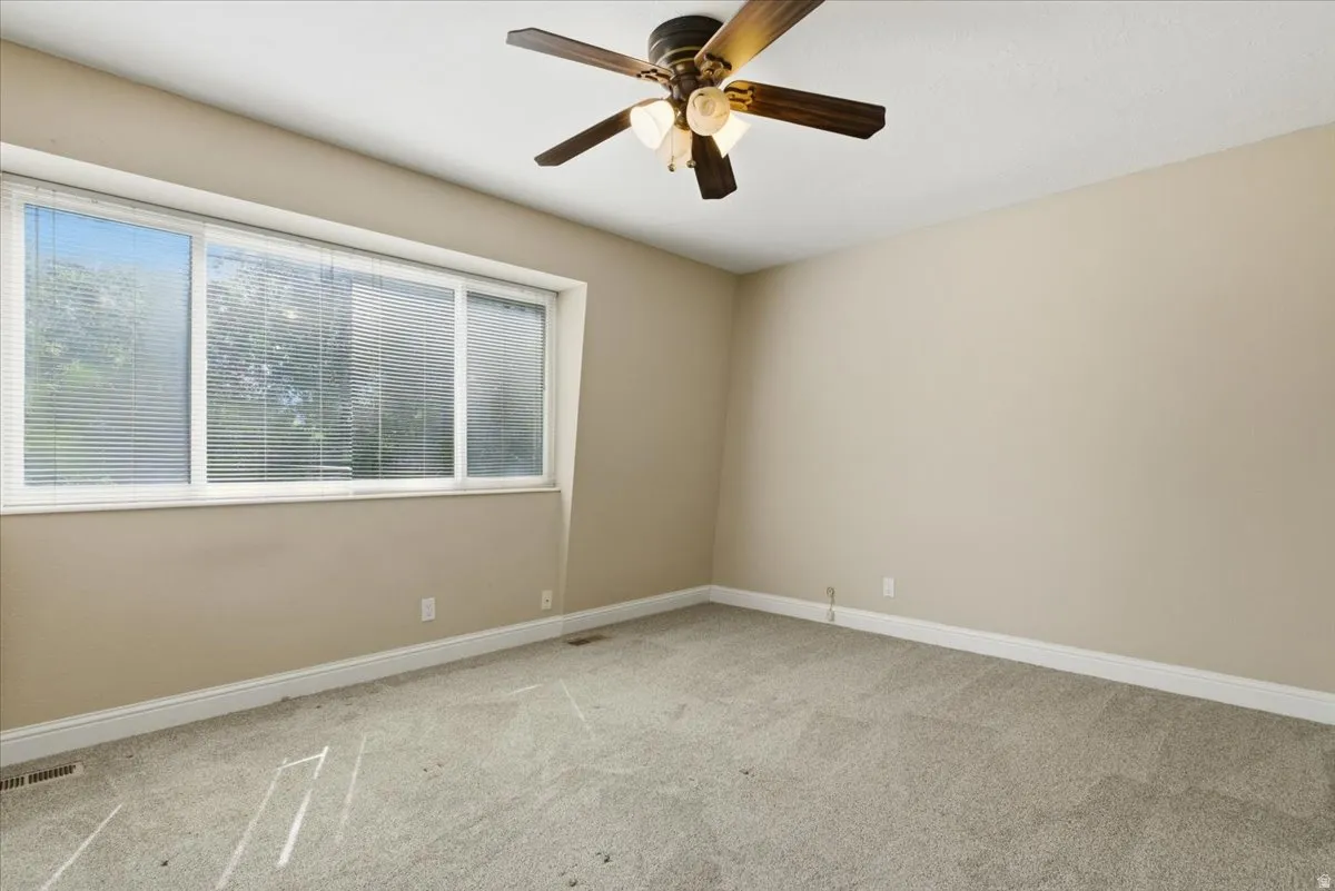 Bright interior space featuring a multi-pane window with horizontal blinds, neutral-toned walls, gray textured carpet, white baseboards, and a ceiling fan with integrated lighting