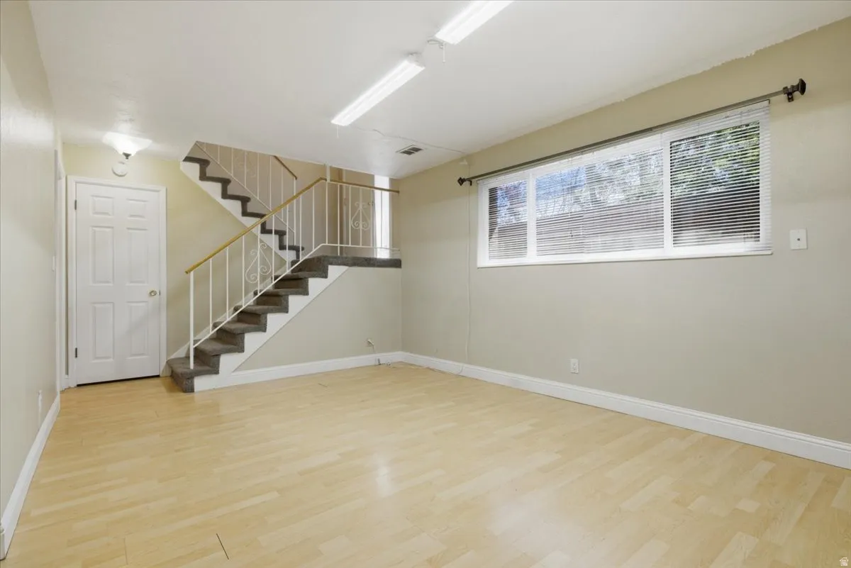 Spacious room featuring light wood-finish flooring, a large window with horizontal blinds, and a staircase with carpeted treads and white metal railings