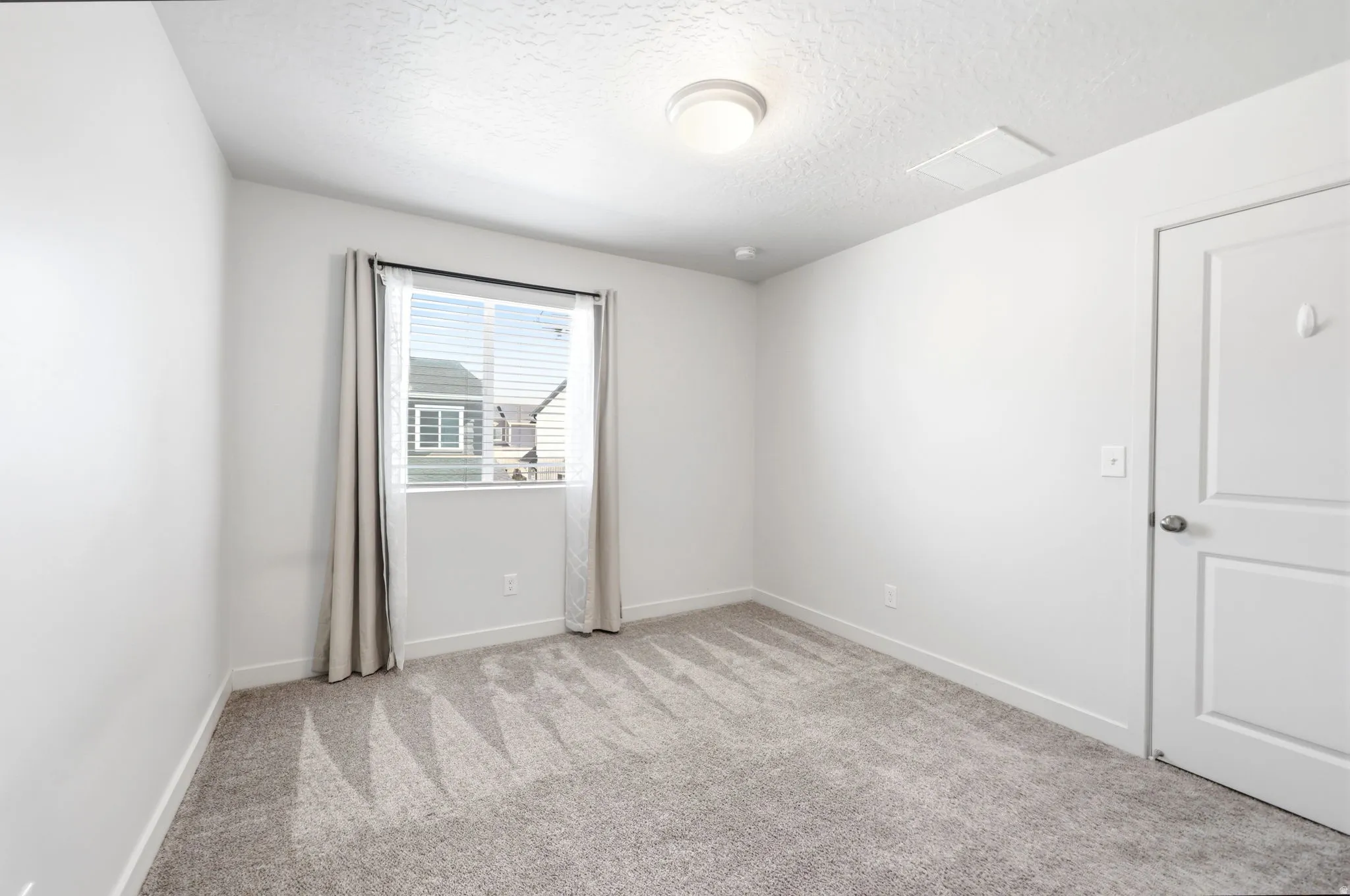Neutral-toned room featuring a single window with blinds, light gray wall-to-wall carpeting, and a flush-mount ceiling light fixture