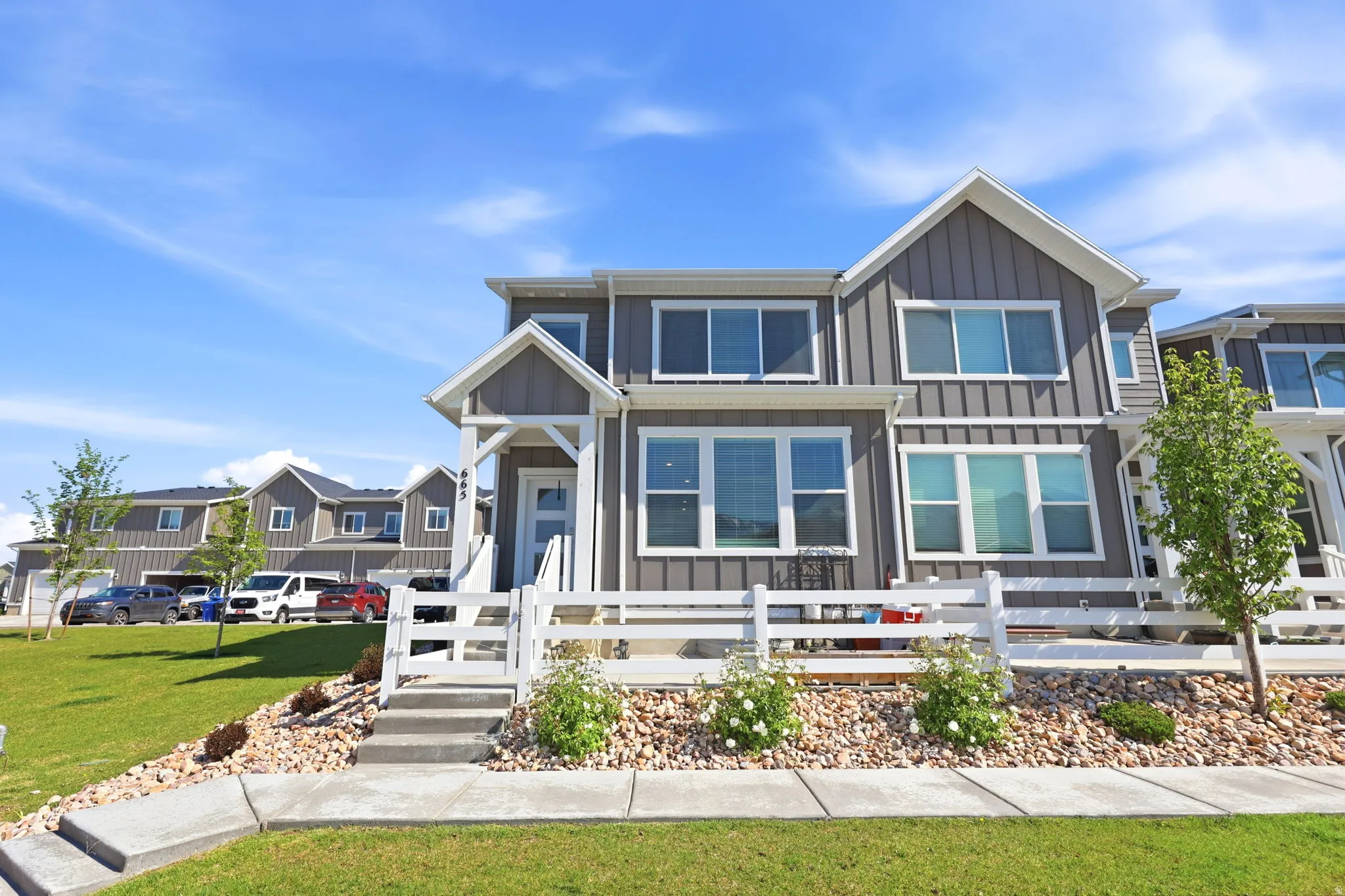 Two-story townhome featuring board and batten siding, multiple windows, a white picket fence, a covered porch, and concrete steps with a landscaped rock bed