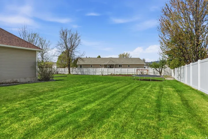 Expansive grassy yard with a white privacy fence, mature trees, a shrub, and beige siding on the adjacent structure