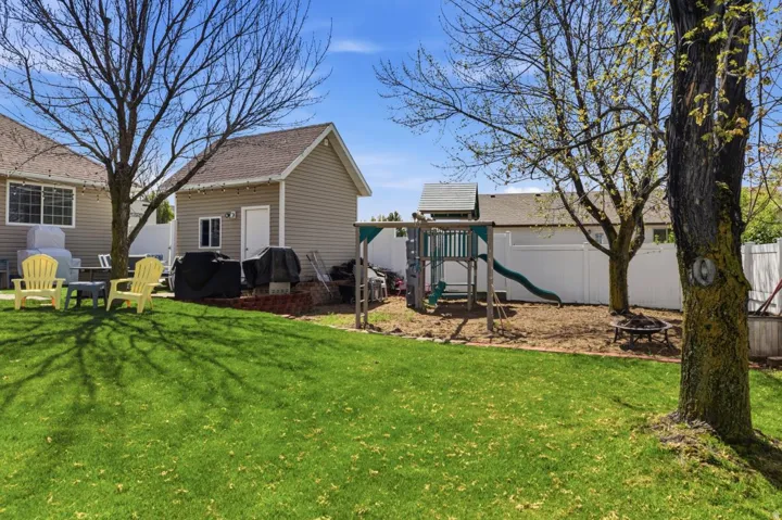 Expansive backyard featuring a manicured lawn, a detached accessory building with tan siding, a white privacy fence, and mature trees providing shade