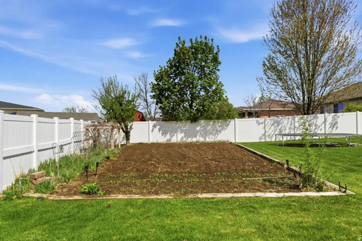 Expansive backyard featuring a large garden bed, lush green lawn, full privacy fencing, mature trees, and a wooden arbor