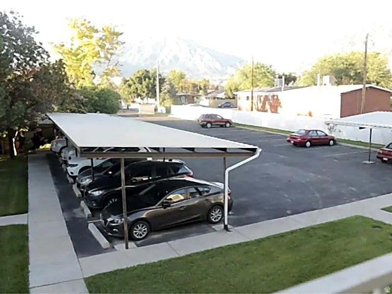 Paved parking lot featuring a covered carport structure, striped parking spaces, and a concrete sidewalk perimeter