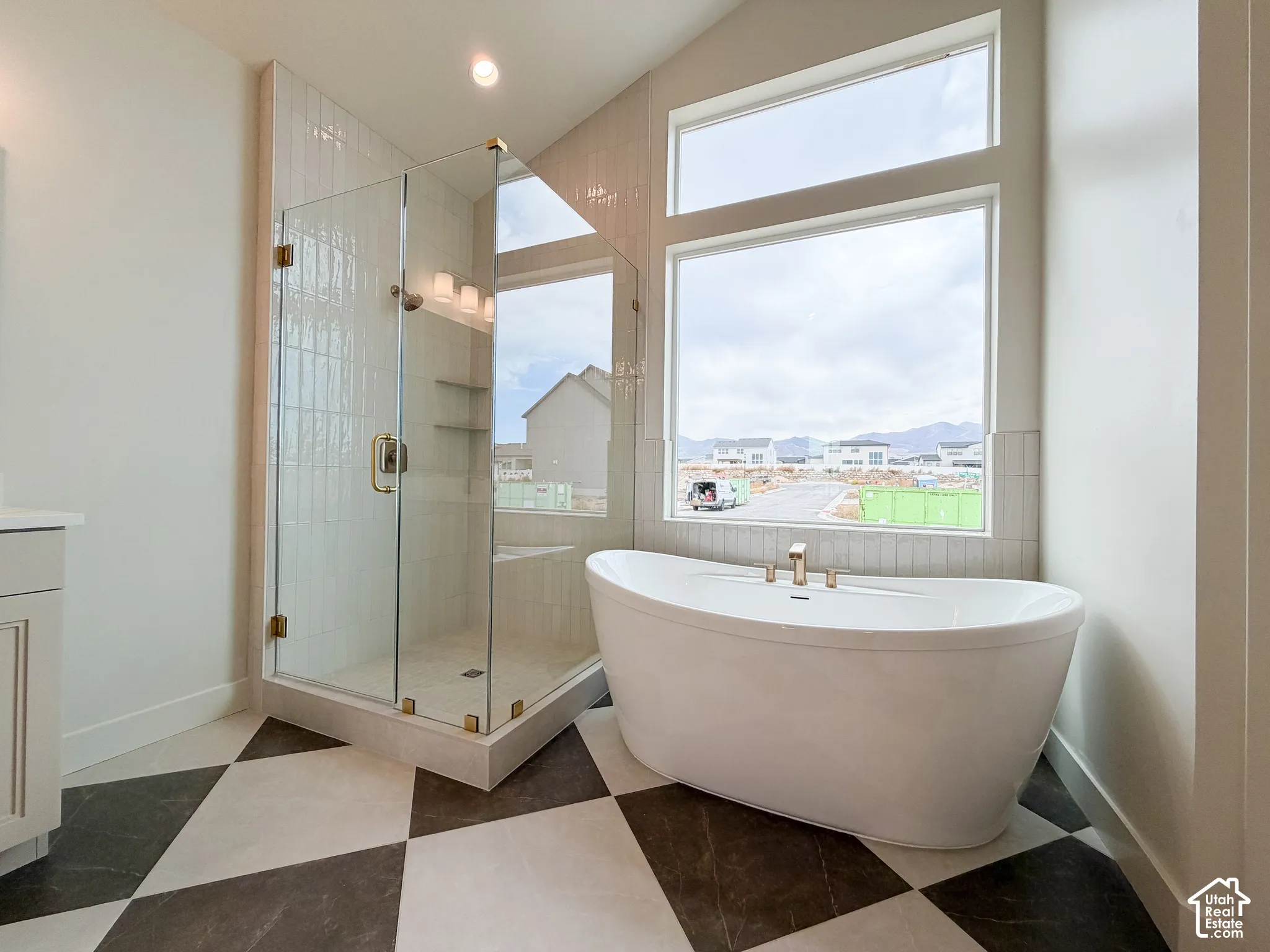 Freestanding soaking tub with gold-finish fixtures, a spacious glass-enclosed shower with tile surround, large windows, and checkerboard floor tiling