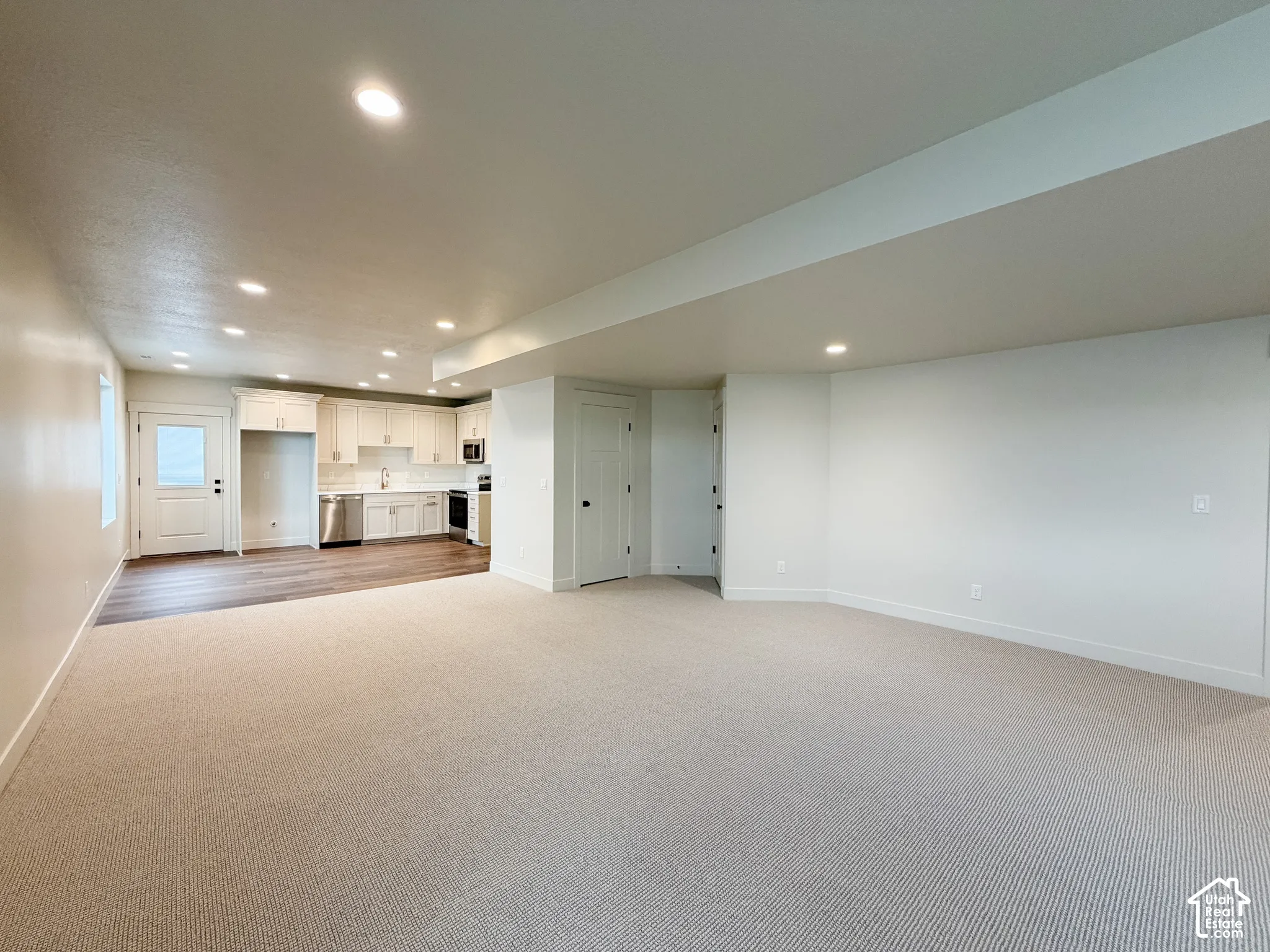 Open-concept living area with plush carpeting, recessed lighting, and a modern kitchen featuring white cabinetry, stainless steel appliances, and wood-finish flooring