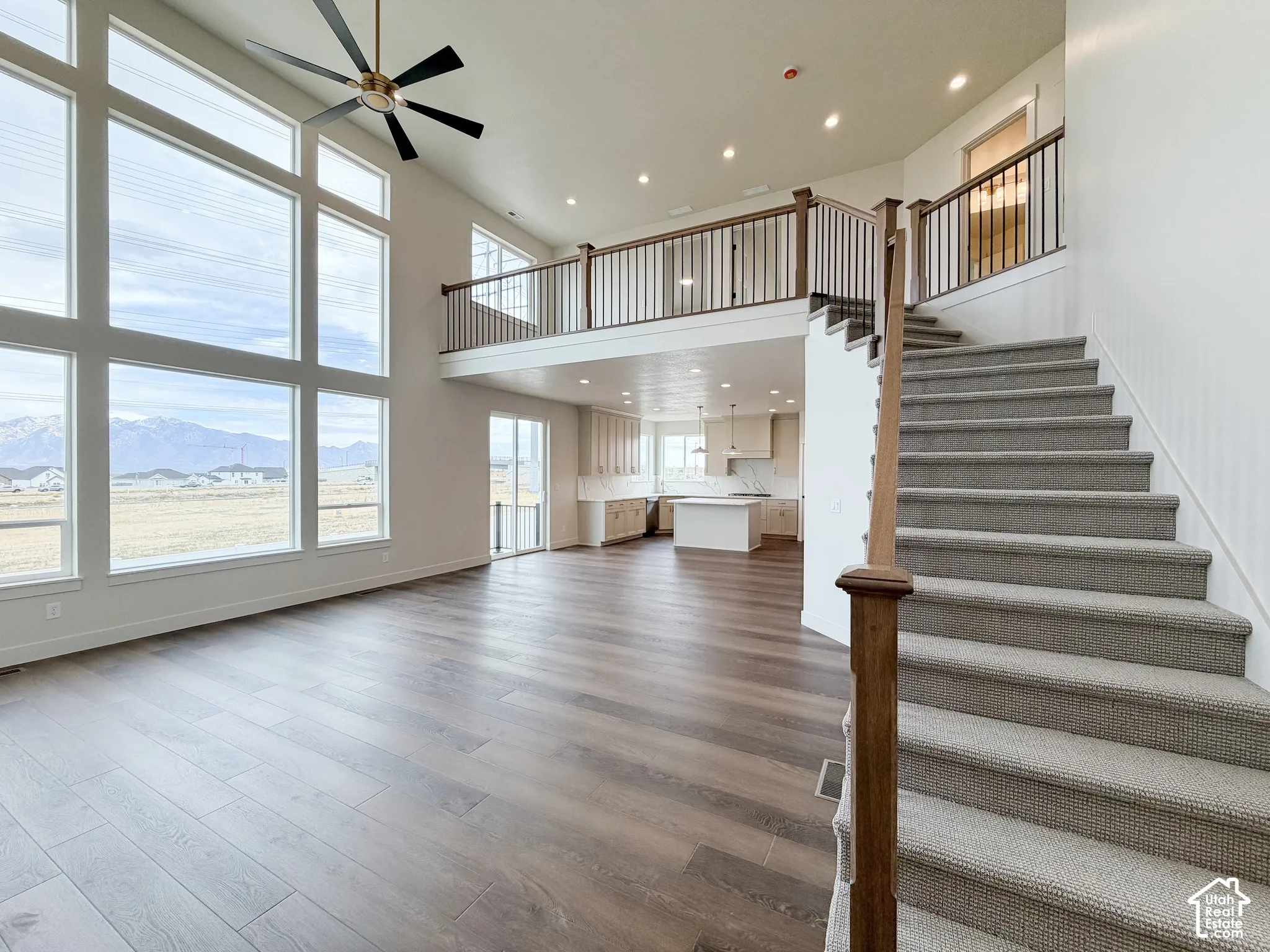 Vaulted ceiling with a ceiling fan, expansive multi-pane windows, wood-finish flooring, staircase with carpeted treads, and an open upper-level landing with iron railing
