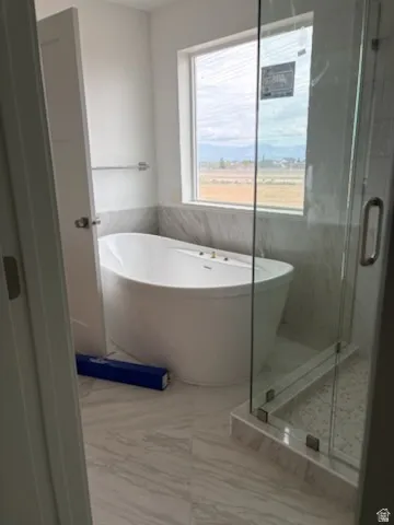 Bathroom featuring a freestanding tub, glass-enclosed shower, large window, and light-toned tile flooring and wainscoting