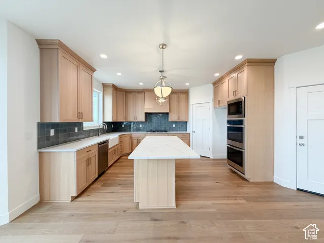 Kitchen featuring wood-finish flooring, light wood cabinetry, a white countertop island, integrated stainless steel appliances, and a dark tile backsplash