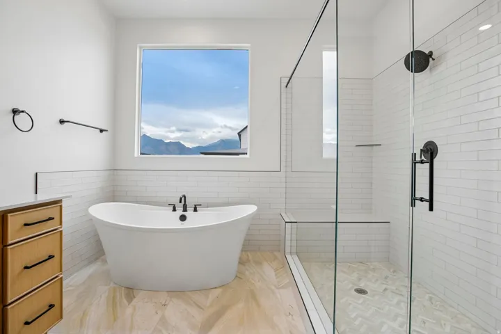 Freestanding soaking tub with black fixtures, walk-in shower with glass enclosure, light wood-finish flooring, and white subway tile accents