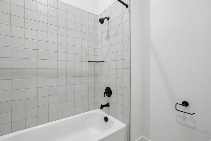 Bathtub with white square wall tiles and matte black fixtures, featuring a glass shower partition and integrated shelving