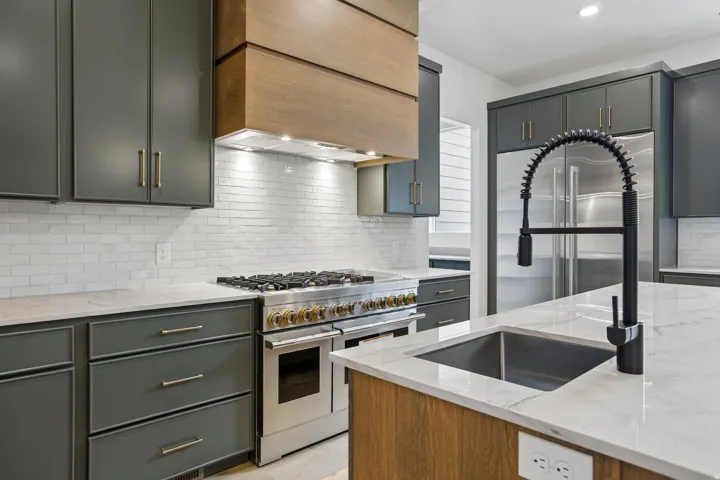 Stainless steel range with gold-tone accents, wood-finish range hood, white subway tile backsplash, dark gray cabinetry with gold-tone hardware, and a kitchen island with an undermount sink and black gooseneck faucet