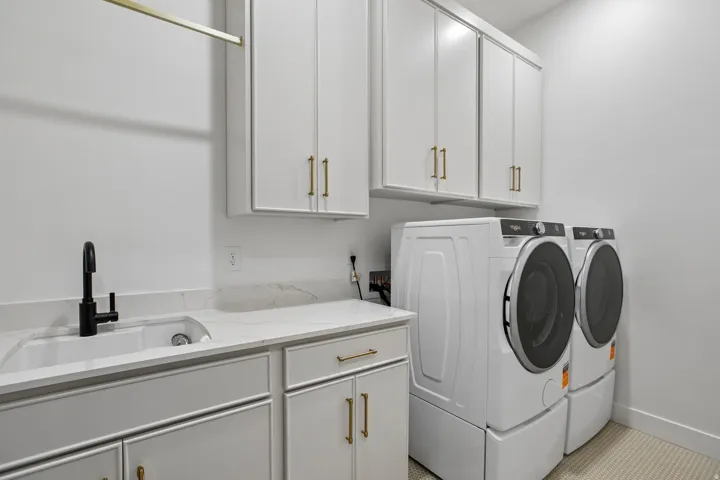 White shaker cabinetry with gold-tone hardware, large basin utility sink with a black matte faucet, light-toned countertop, and integrated washer and dryer units