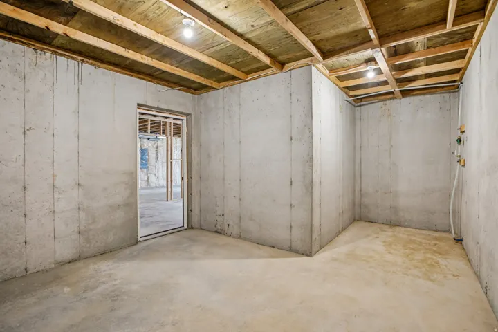 Unfinished basement featuring concrete walls and flooring, exposed wood ceiling joists, and overhead lighting
