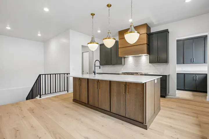 Expansive wood-finish flooring, a large kitchen island with white countertop, three pendant lights, dark cabinetry, and a white tile backsplash