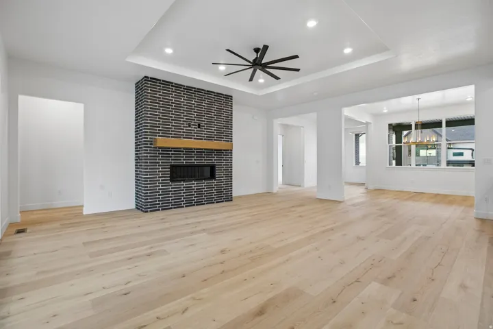 Spacious room featuring wood-finish flooring, a contemporary fireplace with a dark brick facade and wood mantel, a tray ceiling with recessed lighting and a ceiling fan, and large windows
