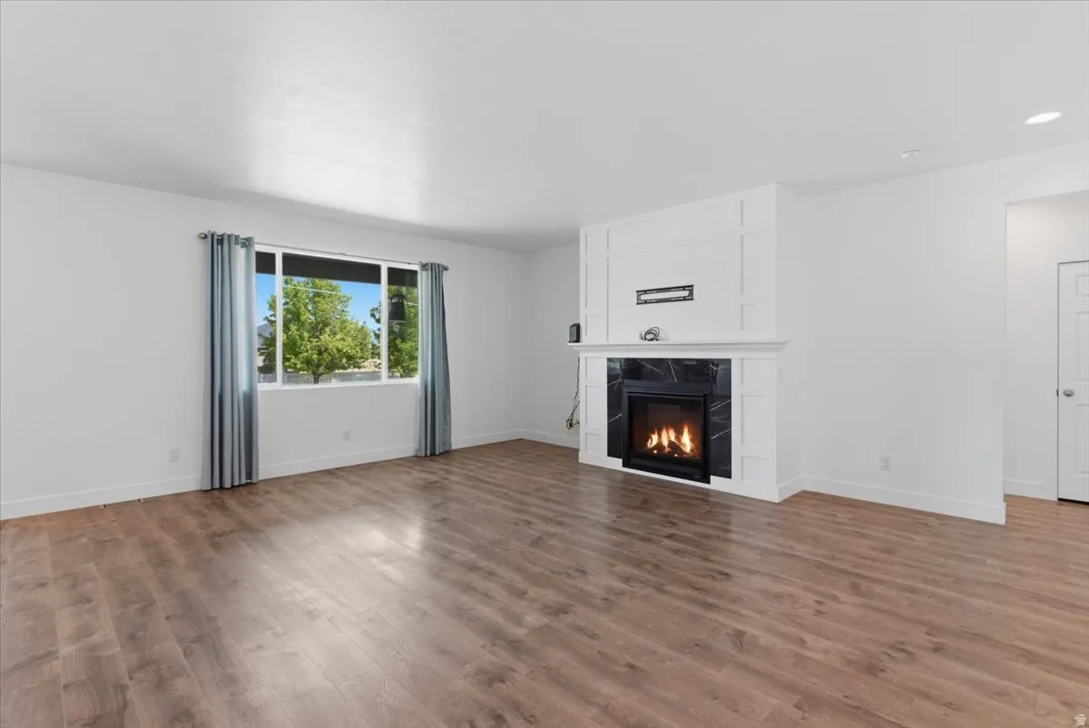 Spacious room featuring wood-finish flooring, a contemporary fireplace with a black stone surround and white mantel, a large window, recessed lighting, and white baseboards