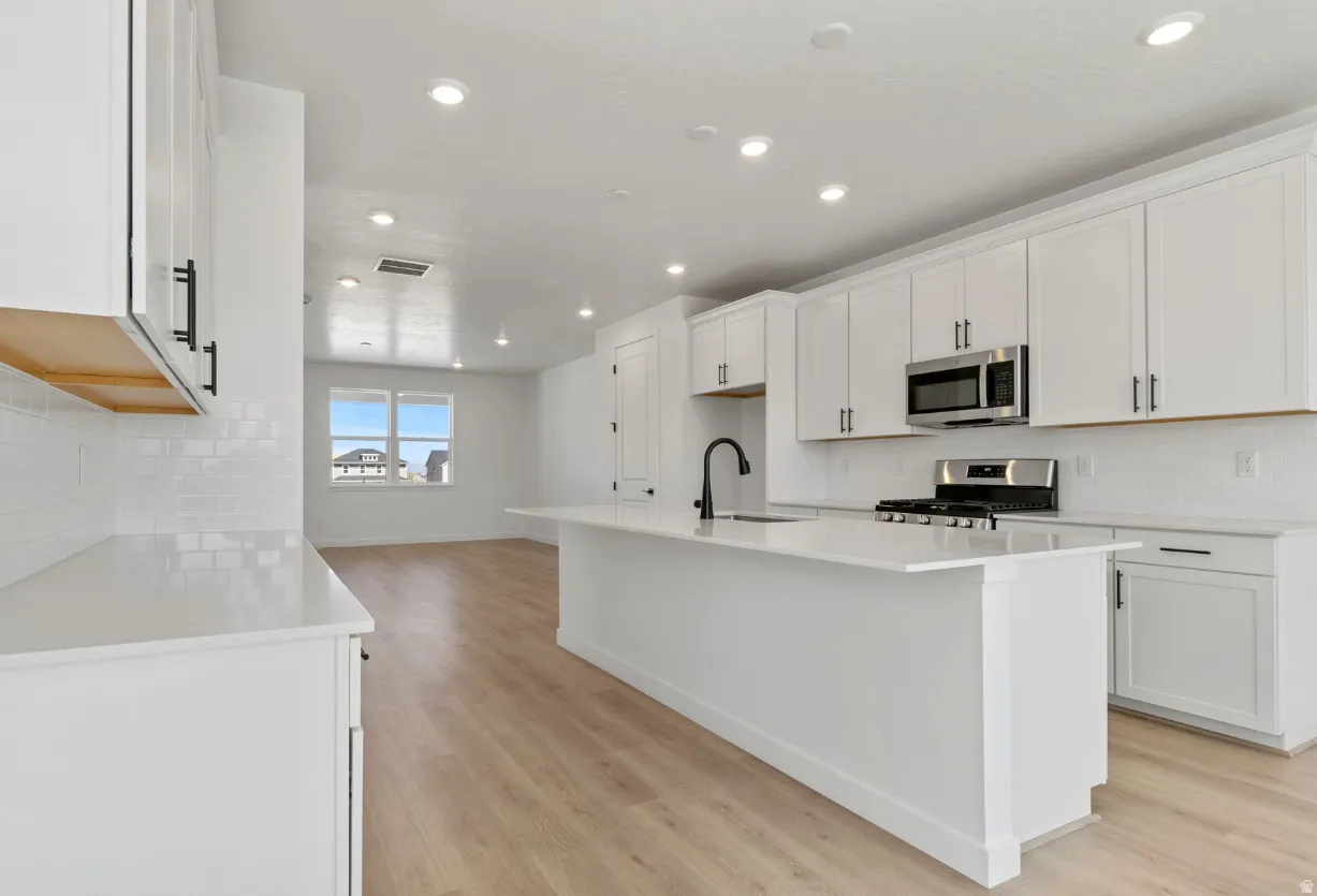 Open-concept kitchen featuring an island with an integrated sink, white cabinetry, stainless steel appliances, and wood-finish flooring