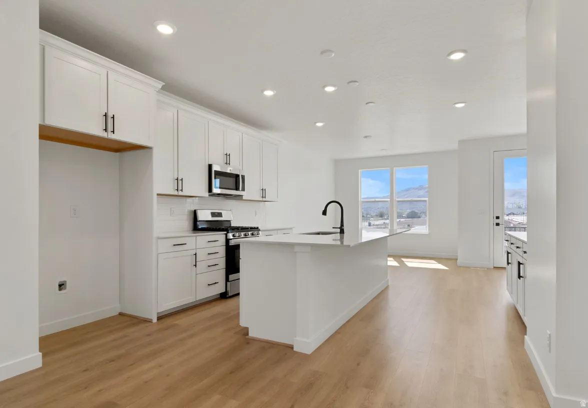 Modern kitchen featuring white cabinetry, a central island with a sink, stainless steel appliances, and wood-finish flooring