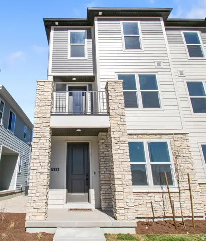 Modern multi-level townhome featuring stone facade accents, light-colored horizontal siding, dark accent siding, a second-story balcony with metal railing, and a dark paneled front door